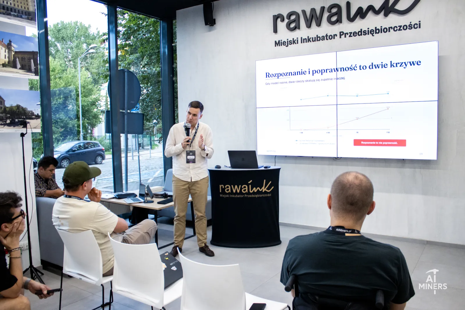 Grzegorz Brzezinka speaking at the front of the RAWA.INK room in Katowice, with the slide &lsquo;Rozpoznanie i poprawność to dwie krzywe&rsquo; projected behind him and the audience in the foreground.